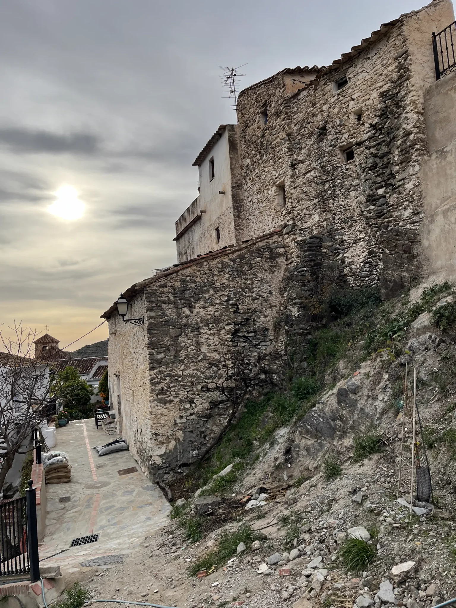 Casa de muros de piedra al atardecer junto a una calle empedrada en pendiente, con el sol tras las nubes
